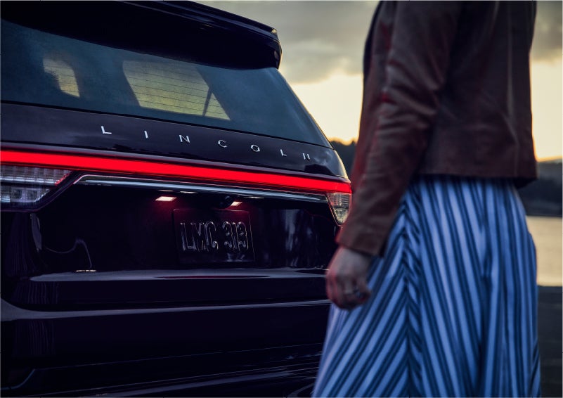 A person is shown near the rear of a 2023 Lincoln Aviator® SUV as the Lincoln Embrace illuminates the rear lights | Fair Oaks Lincoln in Naperville IL