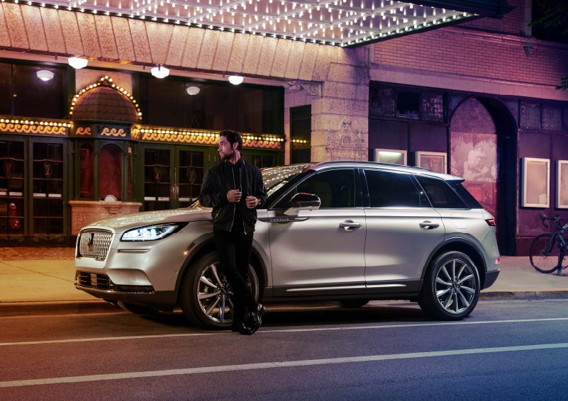 A 2022 Lincoln Corsair SUV is parked outside a theater as the driver relaxes against the frame and lights illuminate the floating roofline and body | Fair Oaks Lincoln in Naperville IL