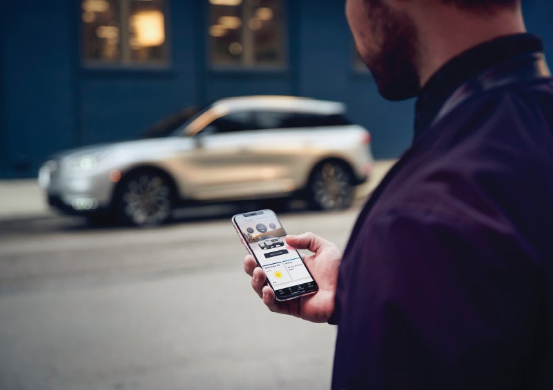 A man is holding his phone looking at the Lincoln Way™ App and we see his 2022 Lincoln Corsair SUV in the background against a blue brick wall | Fair Oaks Lincoln in Naperville IL