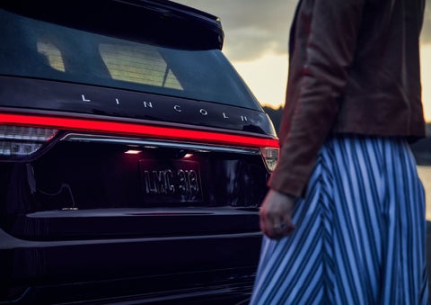 A person is shown near the rear of a 2024 Lincoln Aviator® SUV as the Lincoln Embrace illuminates the rear lights | Fair Oaks Lincoln in Naperville IL
