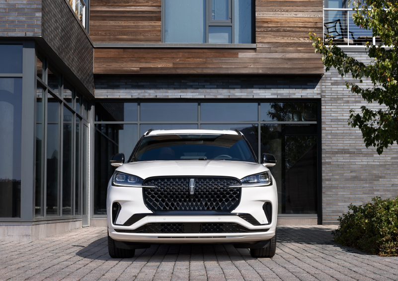 A white 2025 Lincoln Aviator® SUV is shown parked outside a modern building with gray brick and wood accents. | Fair Oaks Lincoln in Naperville IL