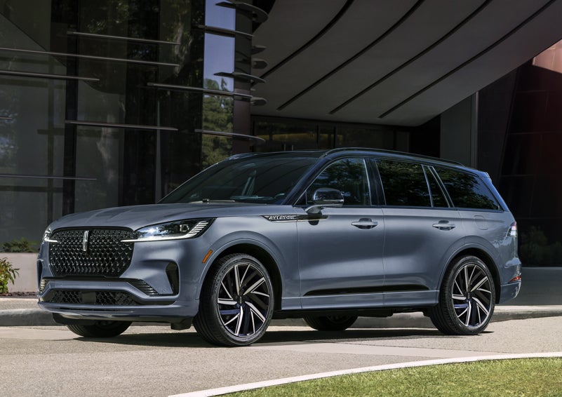 A gray 2025 Lincoln Aviator® SUV is parked outside a modern building showcasing the driver’s side of the vehicle. | Fair Oaks Lincoln in Naperville IL