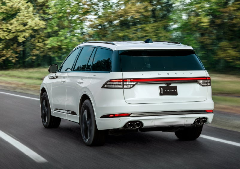A white 2026 Lincoln Aviator® SUV is being driven away down a road lined with colorful trees. | Fair Oaks Lincoln in Naperville IL